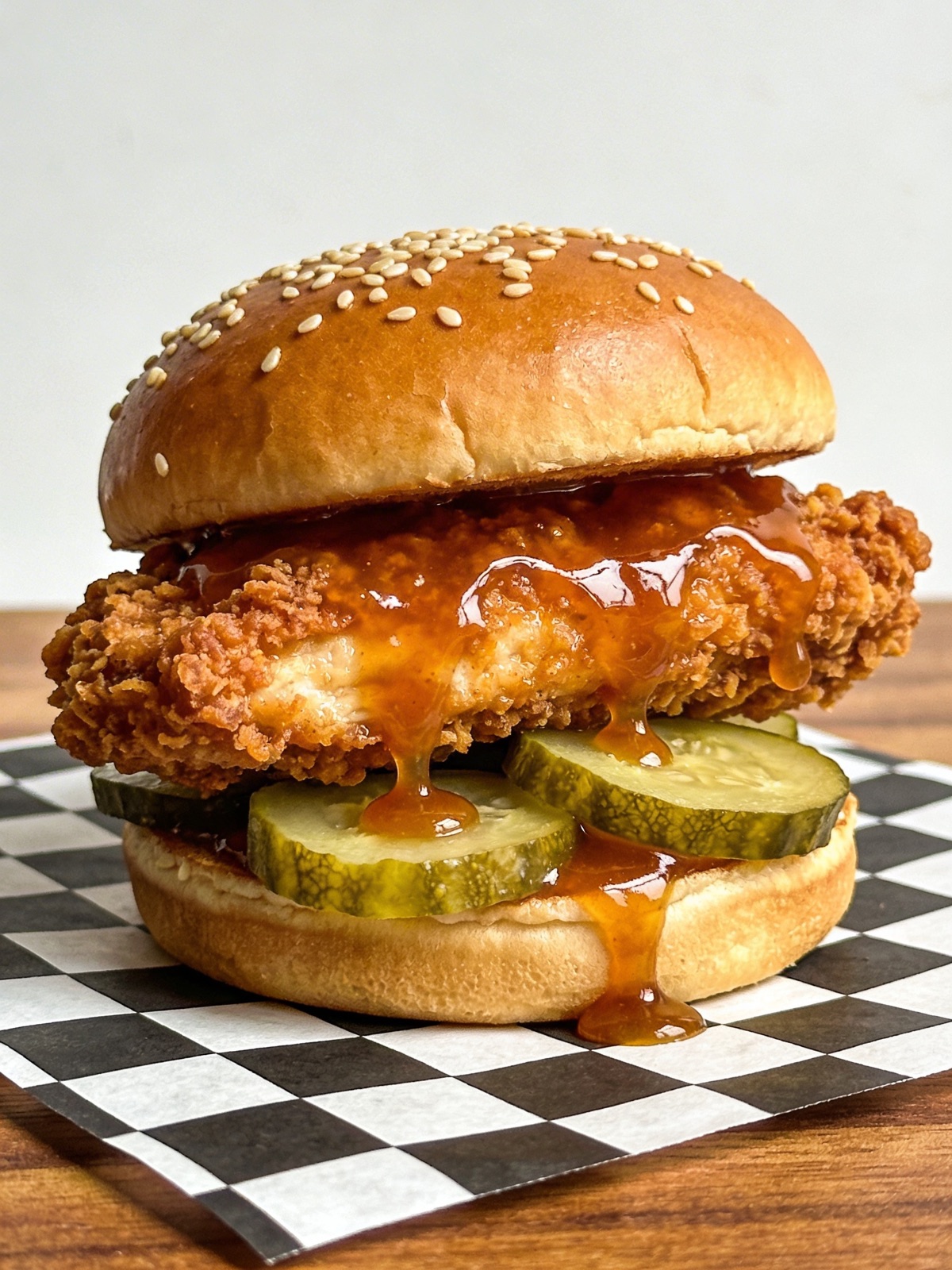 Crispy fried chicken sandwich with hot honey dripping over pickles on a sesame bun, on checkered paper
