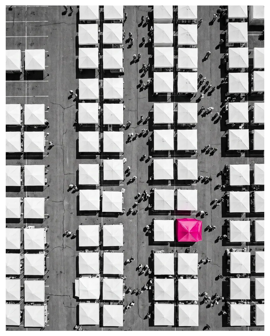 Rows of identical white market tents seen from above, and one hot pink tent among them